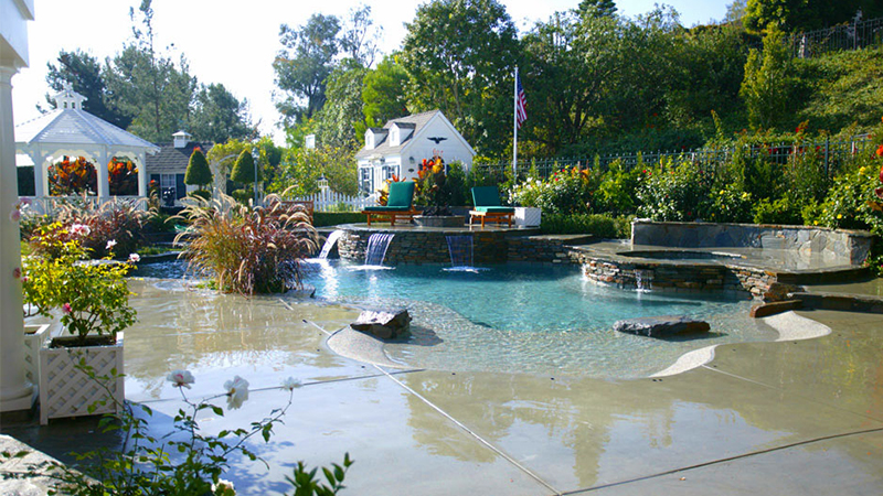 The 2004 NSPI Silver Design Award winner, this Laguna Niguel home was blessed with a generous, open space protected by a steep hillside. Broad, sweeping walkways and stone risers subtly break the islands housing the spa, sun deck and fire pit. A gently sloping beach entry and understated waterfalls suggest a tropical lagoon. The pool area is accented with several varieties of stone, natural brushed concrete and blue Pebblecoat, and finished with Swan landscaping.