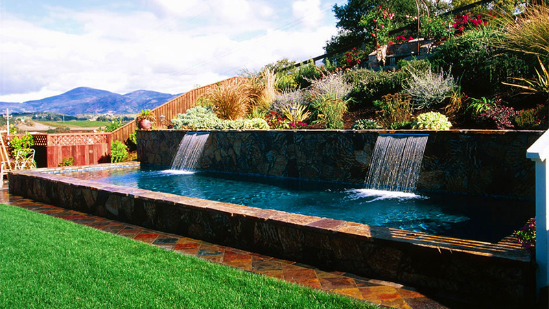 In this Napa Valley property, two dramatic curtains of water reflect the topography of the surrounding hills. They cascade before an abstract mural of stone veneer, down into a geometric basin surfaced in midnight blue. This pool has a timeless quality, as if it had been here for centuries.